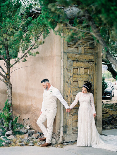 A bride and groom stand side by side, holding hands near a rustic wooden door, embodying the intimate and timeless atmosphere of a luxury wedding.