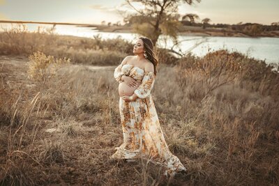 pregnant woman standing in a field during the golden hour. She is pregnant and holding her belly. She is wearing a white and yellow two pice outfit with a flowy skirt. Her hair is long and wavy.