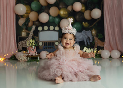 A toddler girl in a large pink tule dress sits in a birthday cap in a studio smiling big