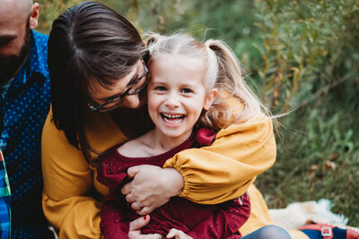 Little girl in red dress smiling as Mom hugs her from behind and looks at her