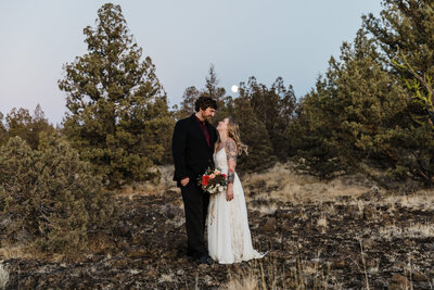 A tattooed bride with long, blond flowing hair wearing a white dress and holding a bouquet of pink, red and white roses looks up at her husband, wearing a black suit and burgundy tie in the desert near Smith Rock as the moon rises above the Juniper trees outside of Bend, Oregon. | Erica Swantek Photography