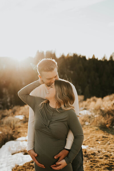 Man holding his pregnant wife's belly from behind as she smiles and touches his neck