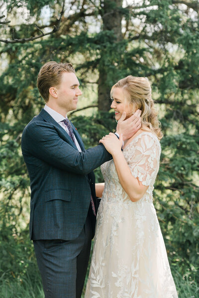 A groom in a suit from Brothers in Edmonton caresses his bride's face during a first look at Millcreek Ravine  during an Edmonton summer wedding