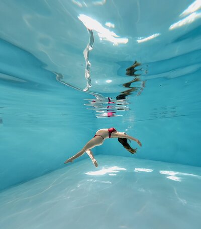 Pregnant woman swimming gracefully underwater in a maternity photoshoot, with soft light reflecting through the clear blue pool.