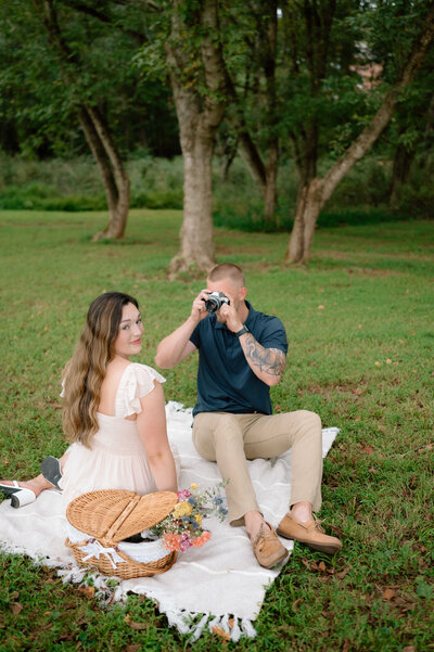 In the midst of a romantic picnic in the park, a couple sits on a white blanket. Beside them, a woven basket holds a collection of wildflowers. The girl turns towards the camera, as the guy captures the enchanting scene with his photography.