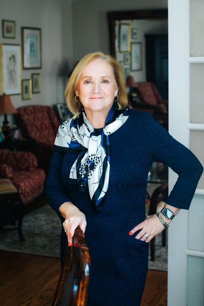 Portrait of a fashionable middle-aged woman in a home she is selling. One hand rests on a dining room chair and the other is on her hip. She's blonde in a navy dress and flowered scarf with a neat red manicure.