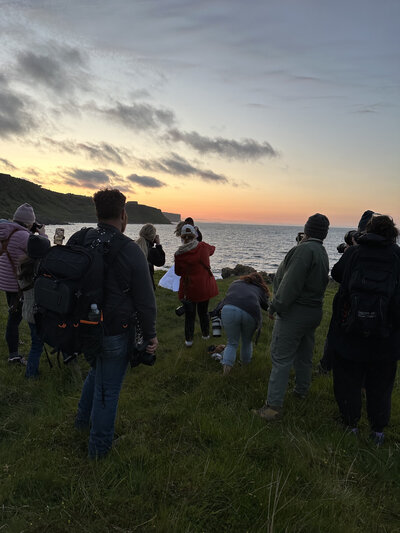 A group enjoys a Colorado family lifestyle session in the wild, standing on a grassy cliff at sunset, overlooking the ocean and capturing playful photos.