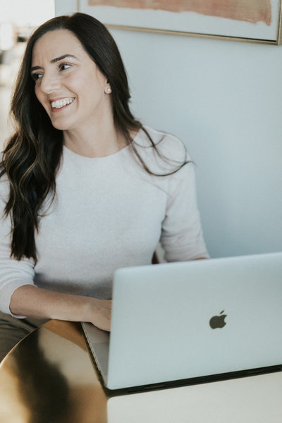 Idit smiles as she sits and types on a laptop. She is an online therapist and marriage counselor in Florida. Learn more from a Florida therapist today.