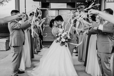 Newlywed couple sharing a kiss while exiting a building, surrounded by guests waving ribbon wands in celebration, in a black and white photograph coordinated by an Iowa wedding planner.