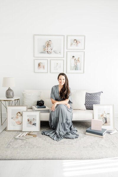 Photographer, Kristin Wood, sits on a white sofa wearing a blue dress surrounded by framed portraits from family photography sessions