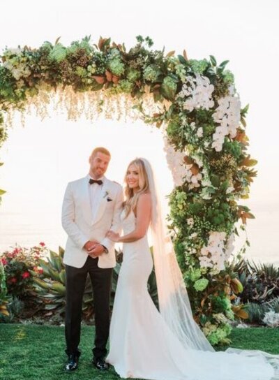 Bride and groom stand smiling under a floral archway during their ceremony