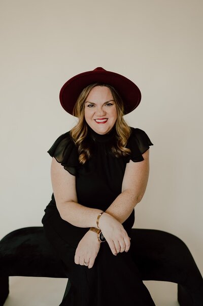 A woman in a black dress and red hat sits smiling on a black bench against a cream background, perfectly capturing the essence of stress-free weddings.