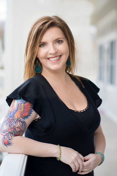 A waist-up headshot of Karen Kahn, of Looking Up Photography. Her colorful right arm tattoo is prominently shown off, and she wears a confident smile, dangly earrings, and a flattering black top.