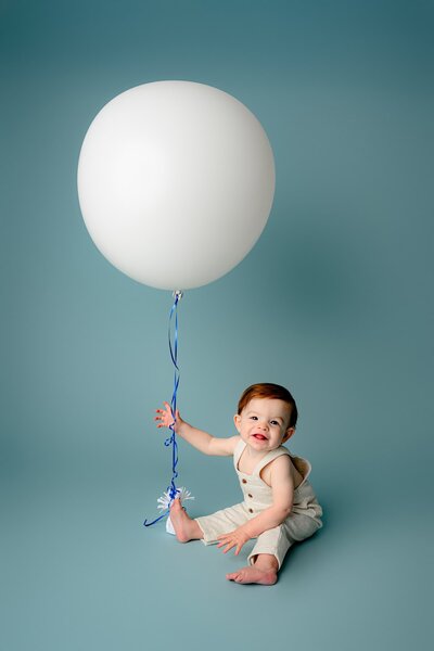 little boy sits on cloth overalls holding a large white balloon on a blue studio background in a charlotte studio set up