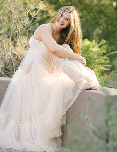 Senior girl sitting on a short wall, in a tulle dress and necklace