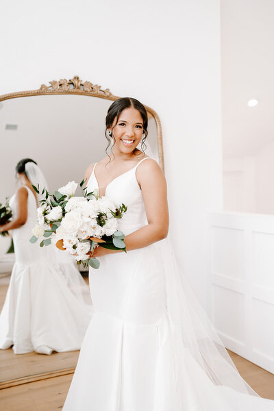 Bride with bouquet standing in front of a grand mirror and smiling