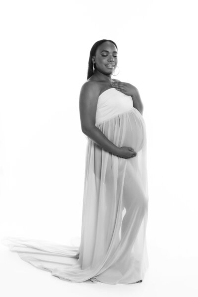 Maternity Portrait of a Black mom with long hair posing with her eyes closed wearing a white dress sheer on the bottom to emphasize her pregnant form, on a white backdrop created in Fort Mill, sc