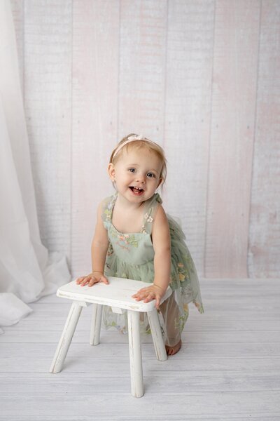 One year old little girl with blonde hair leans against a stool during her birthday photo session in a waxhaw studio