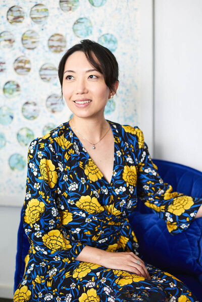 Christen in a flower dress sitting by a patterned wall, looking away from the camera
