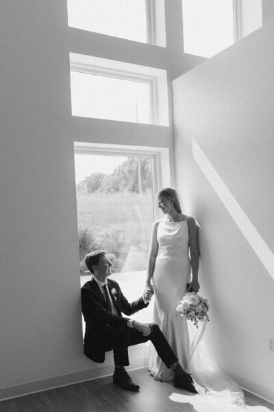 Groom seated on a windowsill, bride standing beside him, hands intertwined, creating a tender moment of connection