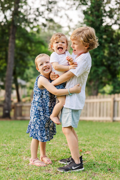 Three children play on a lawn. The older child, a boy in shorts, holds a toddler who is smiling with their tongue out. A girl in a floral dress stands beside them, smiling joyfully. A wooden fence and trees are visible in the background.