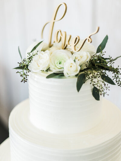 "Love" cake topper with white roses and greenery atop a white wedding cake
