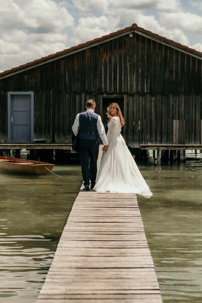 Dieses Hochzeitspaar steht auf einem Steg über dem Wasser vor einer Holzhütte.