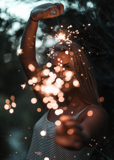 A young blond woman in a white and blue-striped tank top holds out a dazzling sparkler.