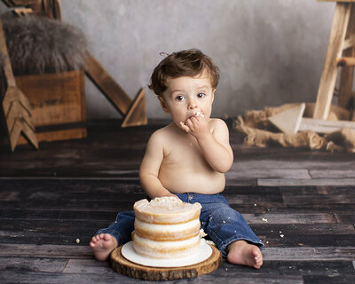 Baby boy eating his First birthday cake while sitting in jeans for his one year portraits in Pittsburgh studio
