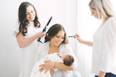 Mom holding her newborn while getting her hair curled by a woman in a white dress and her makeup applied by a woman in a white blouse