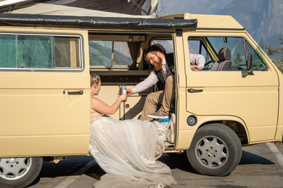 A bride in a white dress and her groom sit inside of a yellow van, drinking a can of beer on their wedding day.