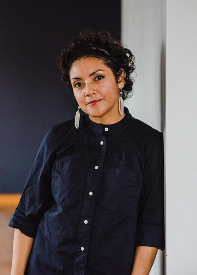 Rosalia, a Latinx woman with curly dark hair and dangling earrings, leans against the wall in a cool black linen shirt buttoned all the way to the top, her arms at her sides, she has an easy-to-talk-to vibe