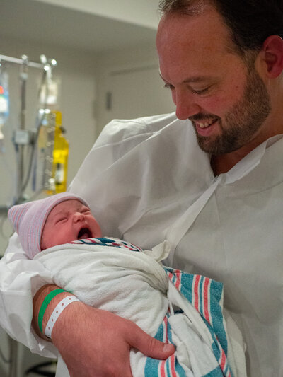 A dad holds his baby while wearing the OR clothing after his wife has gone through a c-section birth.