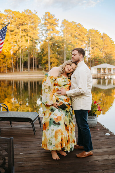 A couple stands on a lakeside dock surrounded by fall foliage. The woman, wearing a flowing floral dress, leans her head on the man's chest as he embraces her tenderly.