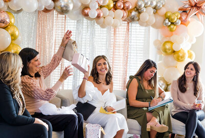 woman opening presents at her bridal shower in a white dress surrounded by friends and family with golden decorations