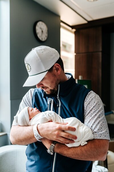 new dad wearing blue vest and ball cap holds brand new baby boy in hospital room in texas