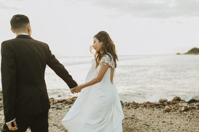 bride and groom holding hands while walking next to ocean