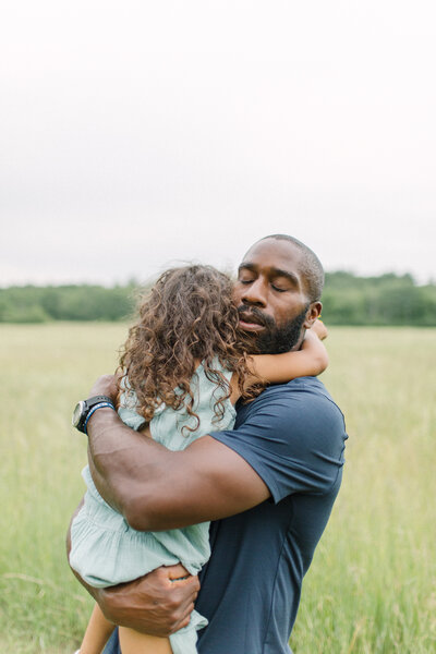 A father is giving his daughter a big hug while closing his eyes standing outside in a beautiful field during photo session with Boston family photographer Corinne Isabelle
