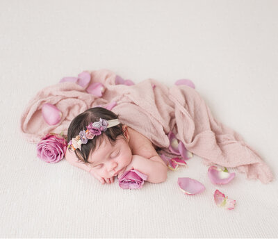 a newborn with rose petals around her and covered by a blanket thats pink and with a white backdrop behind her
