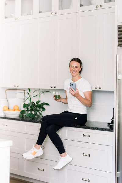 Digital Media Buying Expert Lara Murrant, sitting on kitchen counter with a drink and phone smiling