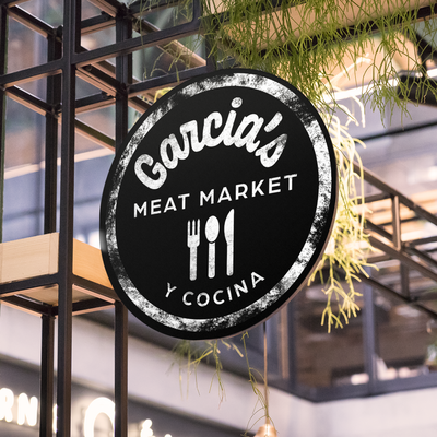 Mockup of logo for Garcia's Meat Market y Cocina on a restuarant sign hanging next to some greenery.