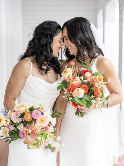 Two brides in white dresses stand close, holding colorful bouquets with pink, orange, and yellow flowers. They are smiling with foreheads touching, creating a joyful, intimate moment. The setting is a bright, airy hallway.