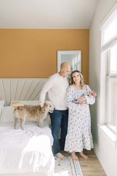 Relaxed husband pets dog standing on bed while wife cradles baby during Austin newborn session.
