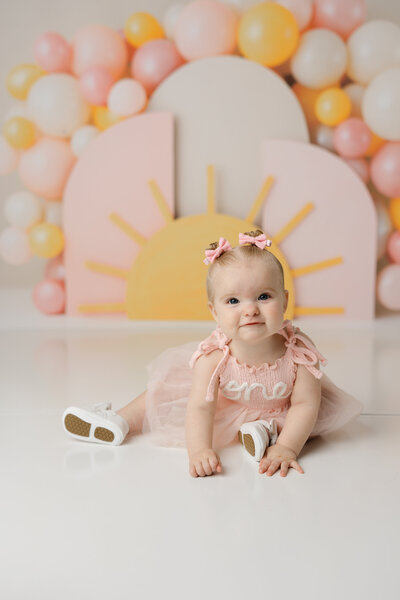 Smiling baby girl in a soft pink dress and matching bows, posing in front of a cheerful sunshine-themed setup.
Photograph by Yucaipa cake smash photographer Kaitlyn Dawn Photography.