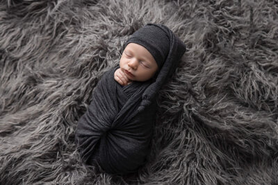 newborn baby wrapped in grey fabric wearing a sleeping cap  asleep on shaggy grey rug