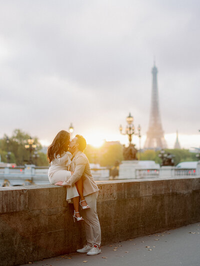 a woman in a white dress sitting on a ledge and a man in a tan suit holding her with the sun setting behind them with the eiffel tower in the distance