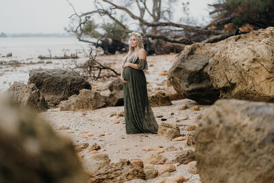 A woman standing amongst the rocks on the beach wearing a green dress and flower crown during her pregnancy photoshoot with Tashina Narelle
