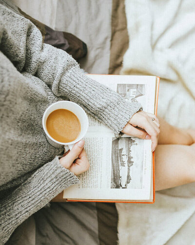 Woman in gray sweater holding a coffee cup and reading a book