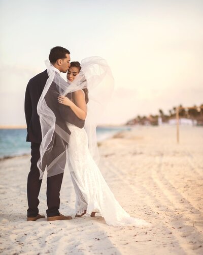 Groom kissing bride on forehead standing on beach after wedding in Cancun