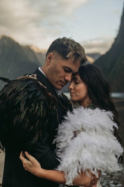 A couple in feathered cloaks embrace outdoors with mountains in the background, capturing the spirit of lifestyle photography for Colorado families.